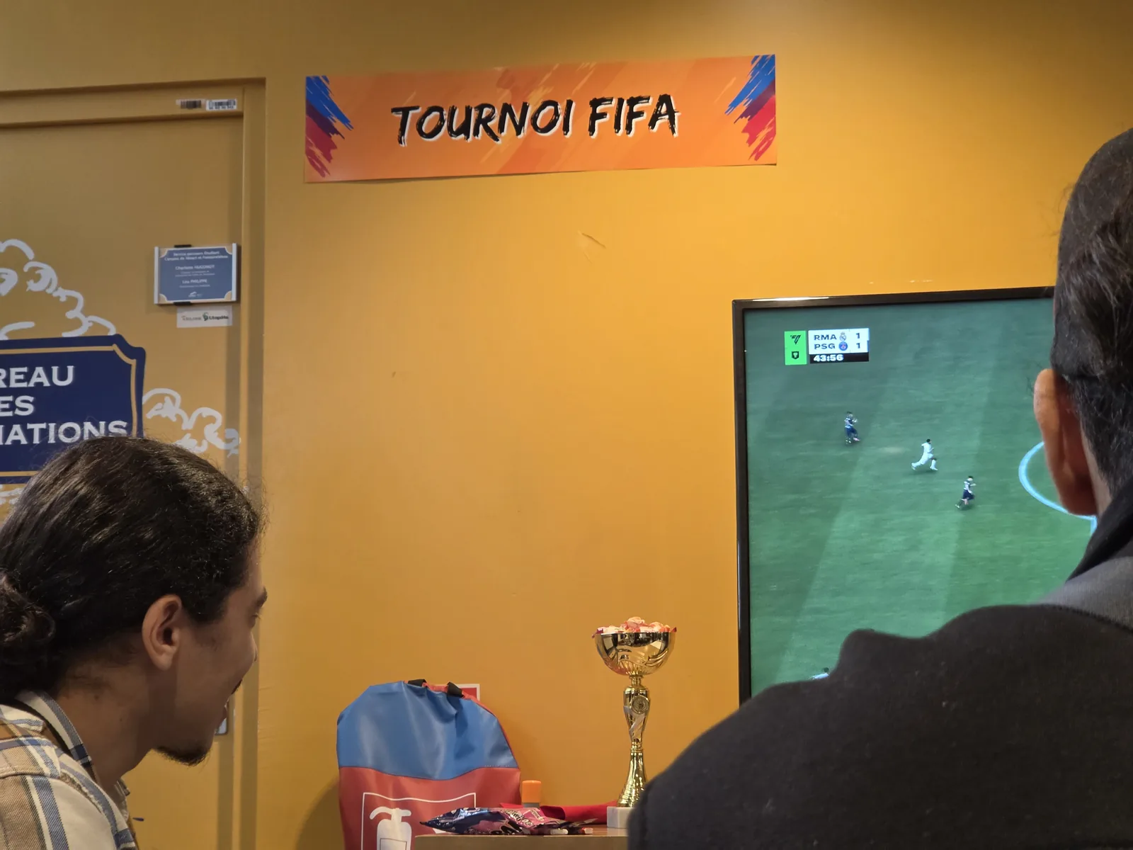 Bandeau TOURNOI FIFA collé sur le mur, deux joueurs concentrés sur leur match avec le trophée et l'écran en arrière-plan