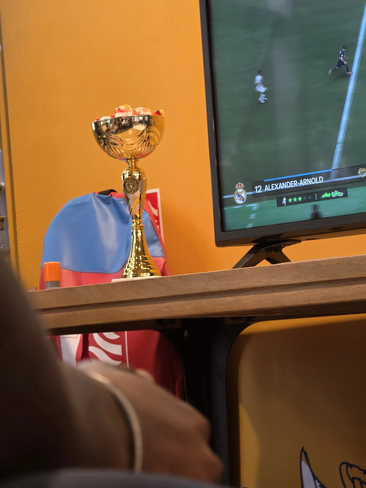 Trophée du Champion FIFA de l'IUT posé à côté d'un écran de jeu, avec la main d'un joueur en action